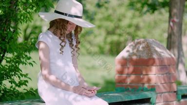 卷曲的女孩白色他衣服板凳上智能手机夏天花园少年女孩浏览移动电话夏天公园绿色植物背景